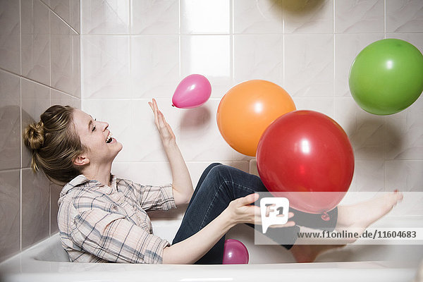 Lachende Frau sitzt in der Badewanne und spielt mit bunten Luftballons