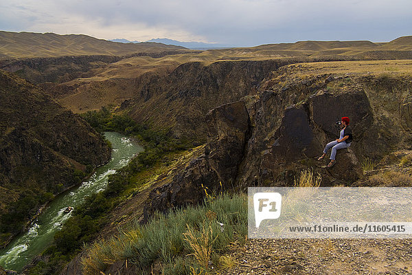 Kaukasische Frau sitzt auf einem Bergfelsen und fotografiert einen Fluss