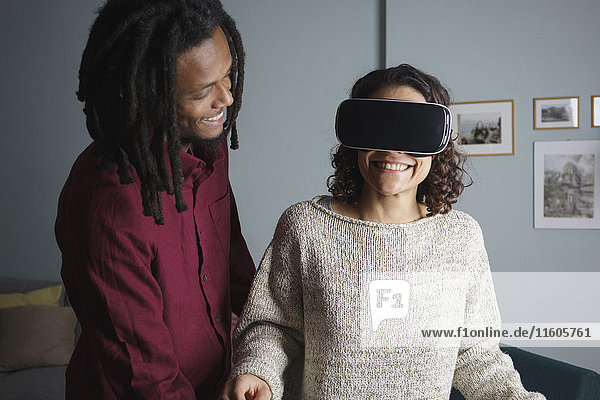 Glücklicher Mann  der eine Frau mit einem Virtual-Reality-Headset im Wohnzimmer ansieht.