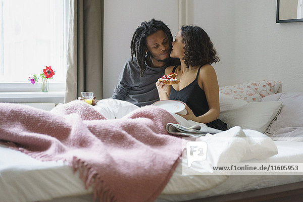 Young woman kissing man while holding pie in bed at home