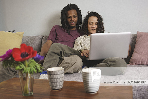 Junges multiethnisches Paar  das zu Hause auf dem Sofa sitzt und einen Laptop benutzt.