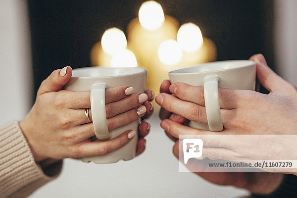 Beschnittenes Bild eines Paares  das Kaffeetassen gegen beleuchtete Kerzen zu Hause hält.