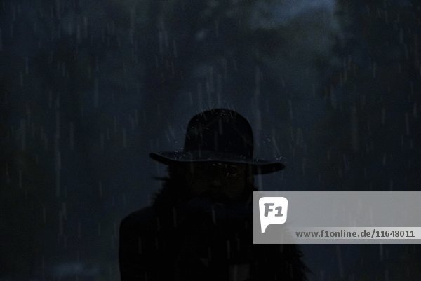 Mysteriöser Mann mit Hut steht im Regensturm mit teilweise entblößtem Gesicht durch Blitzeinschläge