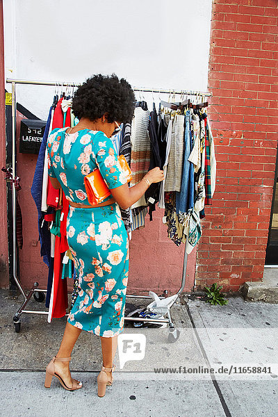 Rückansicht einer jungen weiblichen Mode-Bloggerin mit Afro-Haaren beim Blick auf die Kleiderstange auf dem Bürgersteig  New York  USA