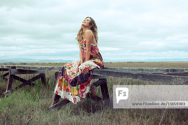 Junge Frau in Boho-Maxi-Kleid auf erhöhtem Holzsteg in Landschaft sitzend
