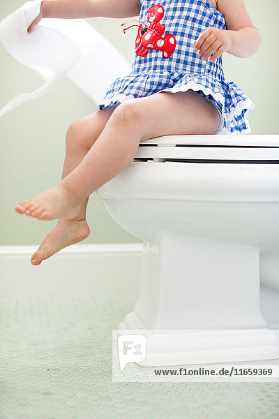 Blick von unten auf den Hals eines weiblichen Kleinkindes  das auf dem Toilettensitz sitzt und Toilettenpapier zieht