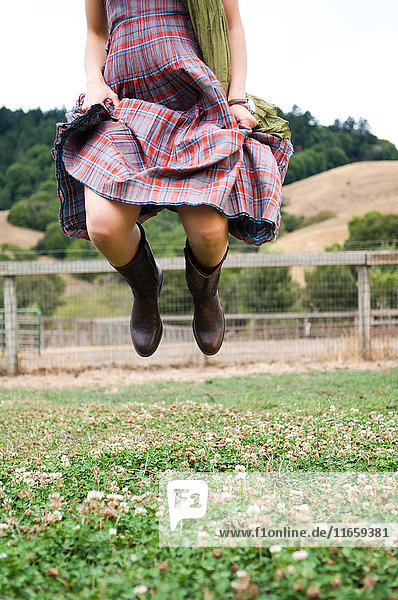Nackenansicht einer Frau in kariertem Kleid  die im Feld in die Luft springt