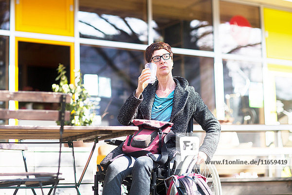 Frau im Rollstuhl  die vor einem Café sitzt und ein kaltes Getränk trinkt