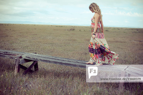 Junge Frau in Boho-Maxi-Kleid geht auf einem erhöhten Holzsteg in der Landschaft