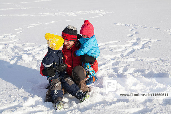 Vater sitzt im Schnee und umarmt zwei Söhne
