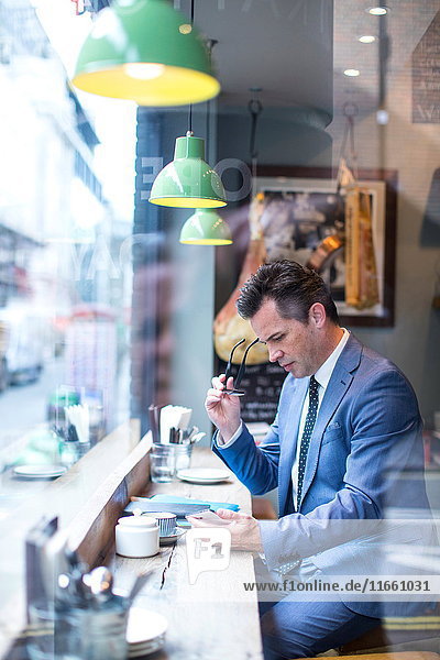 Der reife Geschäftsmann schaut auf das Smartphone im Restaurant-Fenstersitz
