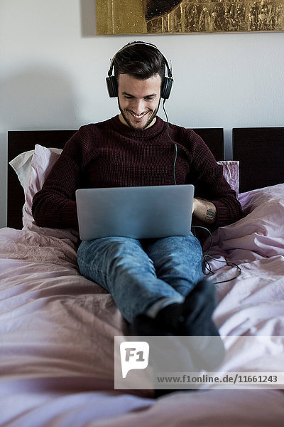 Junger Mann entspannt sich im Bett  benutzt Laptop  trägt Kopfhörer