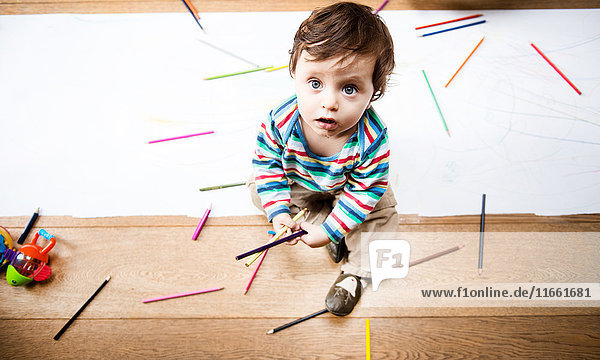 Draufsicht eines auf dem Boden sitzenden männlichen Kleinkindes mit Buntstiften und langem Papier