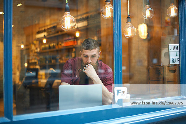 Fensteransicht des jungen Mannes mit Laptop im Cafe