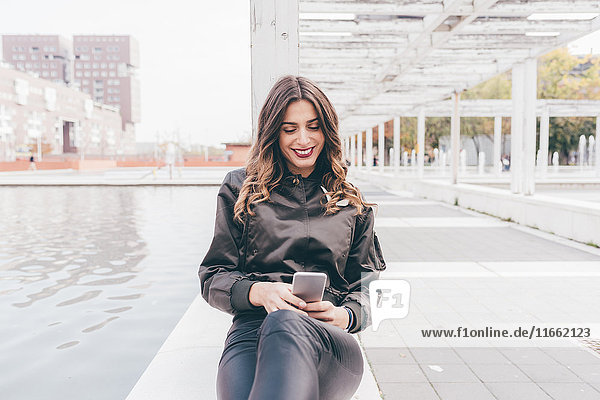 Junge Frau im Freien sitzend  mit Smartphone  lächelnd