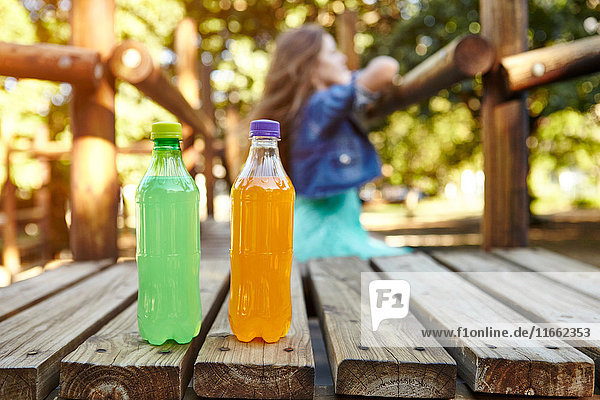 Teenager-Mädchen lehnt auf Klettergerüst im Park  im Vordergrund zwei Flaschen Erfrischungsgetränke