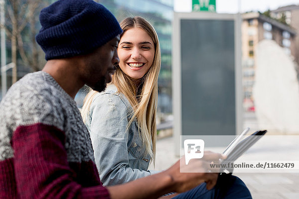 Junger Mann und Frau im Freien sitzend  lächelnd  Mann mit digitaler Tafel