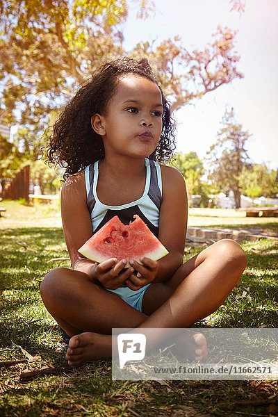 Porträt eines jungen Mädchens  das auf Gras sitzt und eine Wassermelone isst