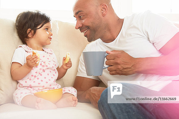 Kleines Mädchen und Vater sitzen auf dem Sofa und essen einen Snack