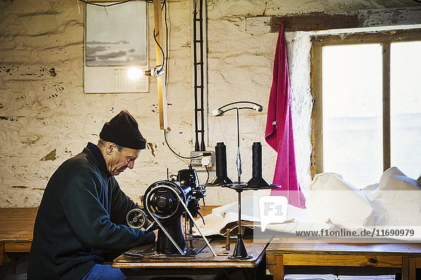 Mann in einer Segelmacherwerkstatt  der mit einer Nähmaschine ein Segel näht.