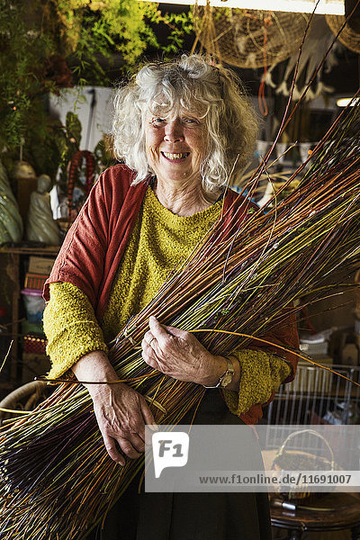 Lächelnde Frau hält ein Weidenbündel in der Werkstatt eines Korbflechters.