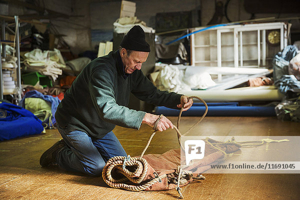 Mann in der Werkstatt eines Segelmachers  der auf dem Boden kniet und ein Seil an einem Segel bindet.