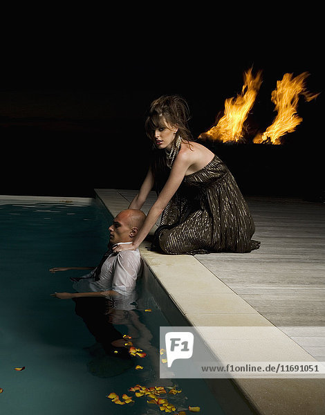 Frau kniet am Rand eines Schwimmbeckens  berührt die Schultern eines Mannes im Schwimmbecken  im Hintergrund Flammen.