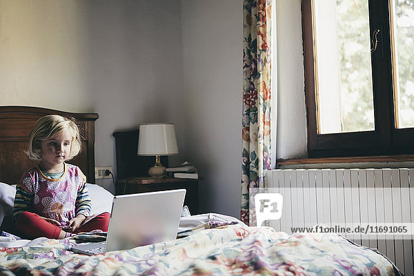 Ein dreijähriges Mädchen sitzt auf einem Bett in einem Hotelzimmer und schaut aufmerksam auf einen Laptop-Computerbildschirm.