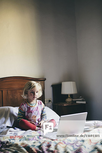 A three year old girl sitting on a bed in a hotel room  looking at a laptop computer screen intently .