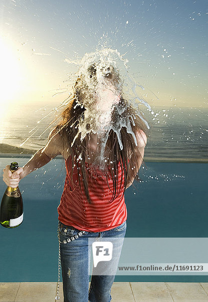 Frau steht vor einem Schwimmbad  hält eine Flasche Champagner in der Hand und spuckt in die Kamera.