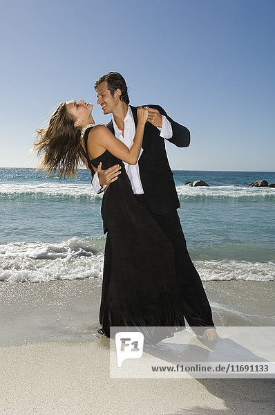 Mann im Anzug tanzt mit einer blonden Frau im schwarzen Abendkleid an einem Sandstrand.