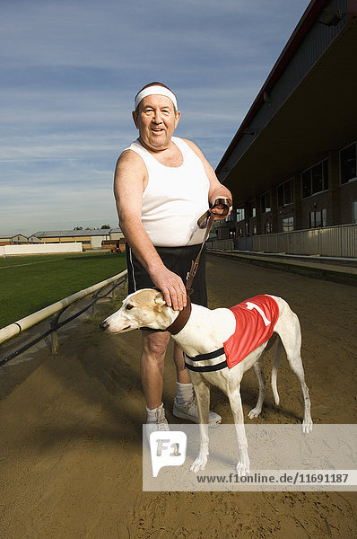 Mann in Sportkleidung steht auf einer Rennbahn  hält einen weißen Windhund mit rotem Lätzchen und der Nummer eins an der Leine.
