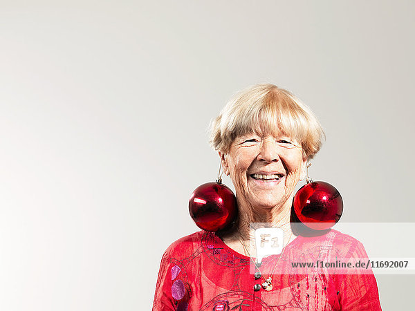 Ältere Frau trägt rote Kugeln als Ohrringe vor weißem Hintergrund