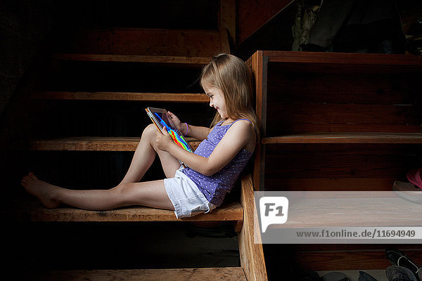 Kleines Mädchen sitzt auf einer Kellertreppe und schaut auf ein digitales Tablett.