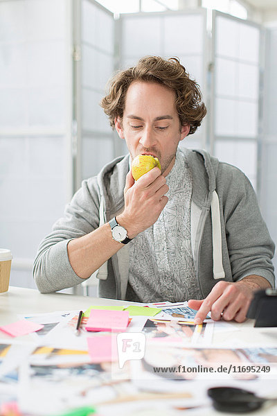 Junger Mann isst Apfel am Schreibtisch im Kreativbüro