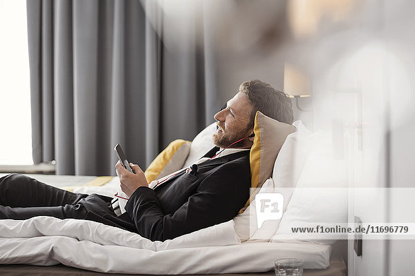 Seitenansicht eines Geschäftsmannes  der Musik hört  während er im Hotelzimmer auf dem Bett liegt.