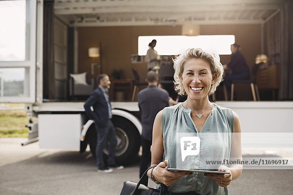Porträt einer glücklichen Geschäftsfrau mit digitalem Tablett und tragbarem Bürowagen auf der Straße im Hintergrund