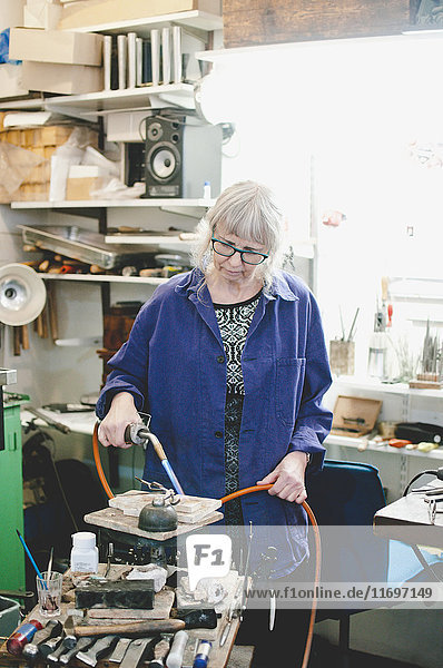 Senior female craftsperson using blowtorch for metalworking in jewelry workshop