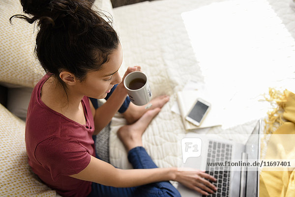 Hispanic woman sitting on bed drinking tea and using laptop