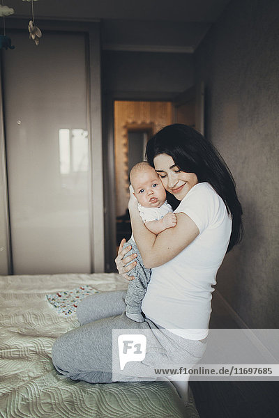 Kaukasische Mutter kniet auf dem Bett und umarmt ihren kleinen Sohn