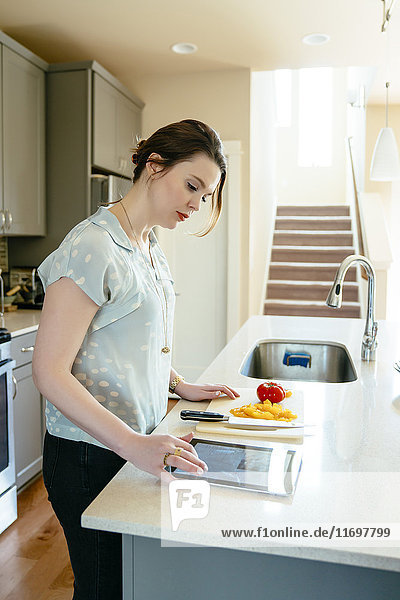 Frau  die ein digitales Tablet benutzt und Lebensmittel in der heimischen Küche zerkleinert