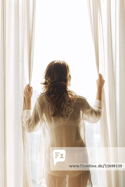 Thai woman wearing sheer clothing opening curtains at window
