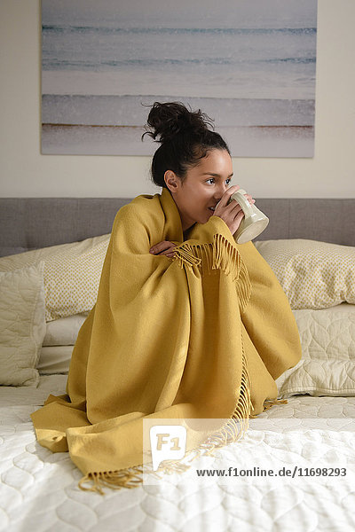 Hispanische Frau auf dem Bett sitzend,  in eine Decke eingewickelt,  Kaffee trinkend