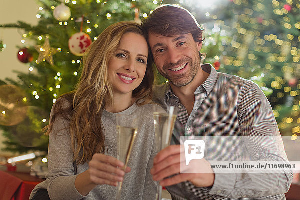Portrait lächelndes Paar stößt mit Sektflöten vor einem Weihnachtsbaum an