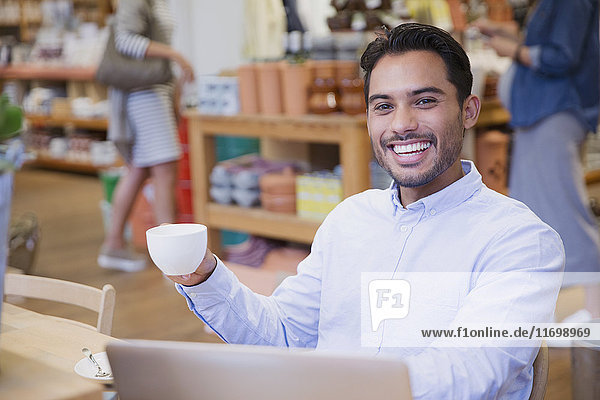 Porträt eines begeisterten jungen Mannes  der in einem Café am Laptop Kaffee trinkt