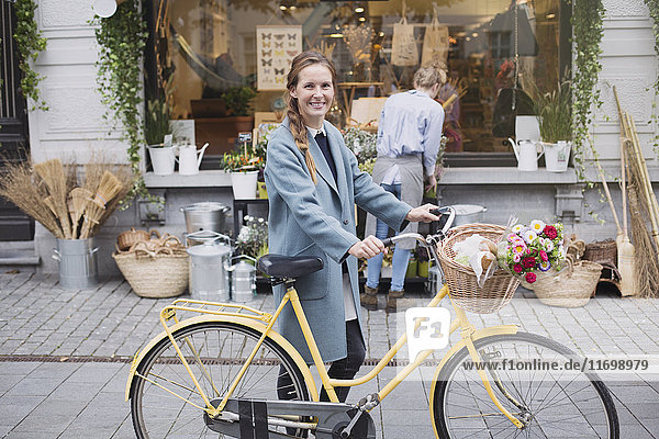 Portrait smiling woman walking bicycle with flowers in basket outside storefront
