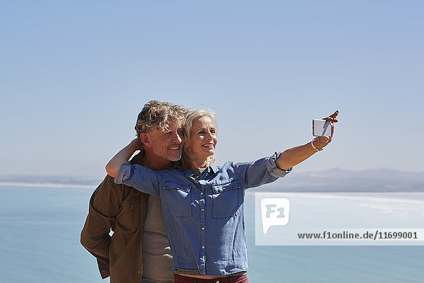 Affectionate senior couple taking selfie overlooking sunny ocean view