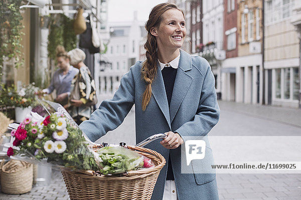 Smiling woman walking bicycle with flowers in basket on city street