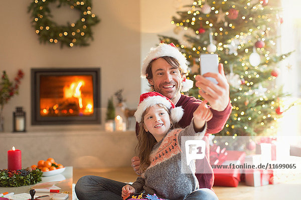 Vater und Tochter mit Weihnachtsmannmützen machen ein Selfie mit Fotohandy im Weihnachtswohnzimmer