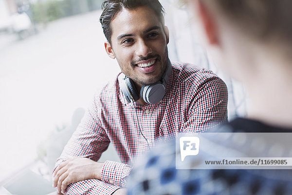 Lächelnder junger Mann mit Kopfhörern im Gespräch mit dem Fenster eines Cafés